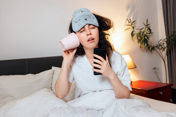 Portrait of sleeping young woman wearing sleep mask call with cup and drink with phone while...