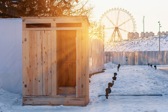 Wooden toilet with open door on a snowy construction site in the winter morning sun. Ferris wheel in the background