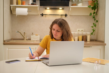 A woman with a laptop is writing with a pen in a notebook while sitting at a table in a home kitchen. An adult female businesswoman works from home, a remote office
