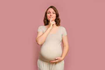 A pregnant woman with her finger on her lips on a pink studio background. Pregnancy in a woman with...