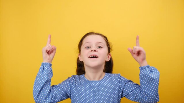 Portrait of happy little girl child 8-9 years old in blue dress point index finger overhead on copy space mockup, posing isolated on yellow color background wall in studio. Childhood lifestyle concept