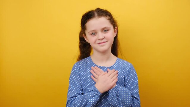 Portrait of kind pretty smiling little girl kid 8-9 years old in blue dress put folded hands on heart sign, posing isolated over yellow color background wall in studio. Childhood lifestyle concept