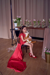 a girl in a red dress with a long train sits hugging a toy meerkat surrounded by tulips