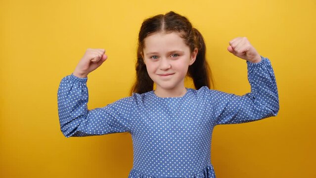 Portrait of strong sporty fitness little girl kid 8-9 years old wearing blue dress showing biceps muscles on hands, posing isolated over yellow color background wall in studio. Childhood concept