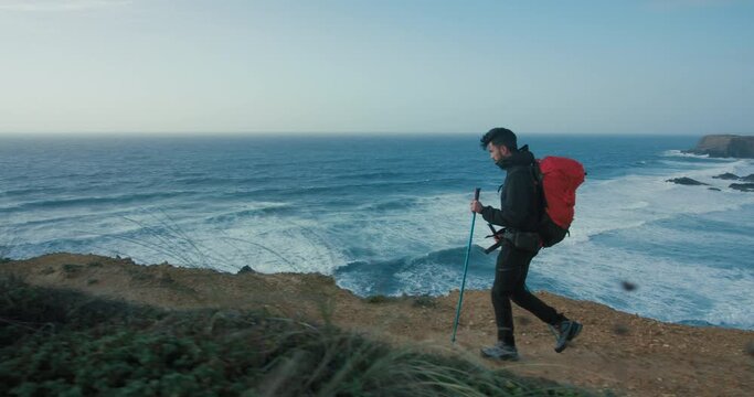 Side shot of young male hiker walk with red backpack and trekking poles alongside ocean. Adventure search and outdoors lifestyle. Backpacking Journey or modern outdoor commercial or advertisement