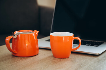 Orange cup and teapot on the background of a laptop on the desktop. Tea break at work. Remote work at home office with tea, cozy workplace.