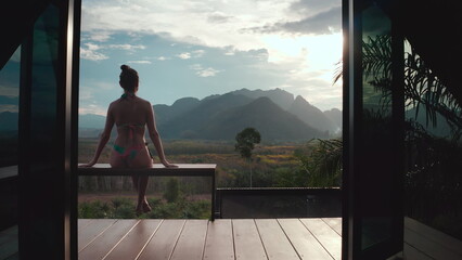 Woman enjoy tropical villa view sitting on terrace. Exotic lonely bungalow in jungles, mountains, forest, sunset. Female tourist on summer vacation. Travel holiday concept. Beautiful wild nature scene