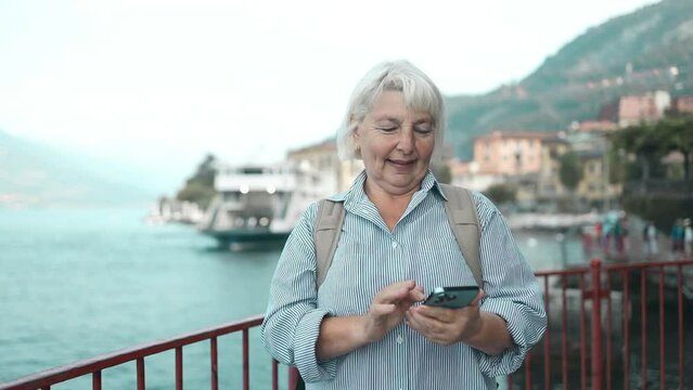Senior 60s woman traveler using smart phone while standing on quay against lake Como, Italy. Female looking at phone screen. High quality FullHD footage