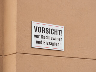 Sign a on a building warning against roof avalanche and icicles in German language. People should be aware during the winter season not to stay next to the wall in the danger zone.