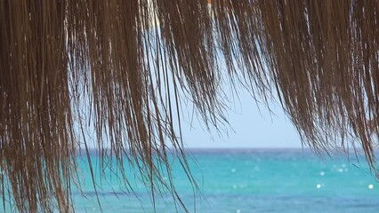 Detail of dry hay hanging from rustic roof eaves waving in summer breeze with turquoise water of Mediterranean sea in background, enjoy shade and nature scenic view on the beach