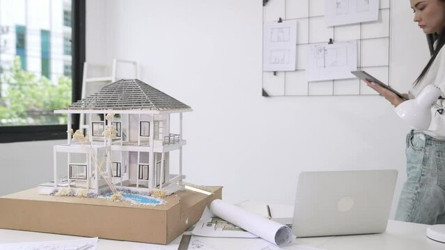 Beautiful caucasian architect focus on using tablet to check house model on meeting table with blueprint, house model and architectural document scatter around at meeting room. Immaculate.