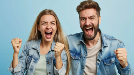 man and a woman are both cheering excitedly with their fists raised, wearing denim jackets, against a blue background