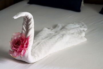 Artistic towel swan with pink rose on bed. Swan towel figure with floral touch on white linen. Pink-flowered towel swan on hotel bedroom bed. Hotel room decoration. Leisure and holidays concept