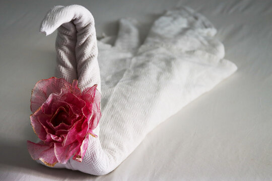 Artistic towel swan with pink rose on bed. Swan towel figure with floral touch on white linen. Pink-flowered towel swan on hotel bedroom bed. Hotel room decoration. Leisure and holidays concept