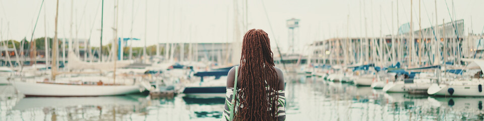 Woman with African braids wearing top looks at the yachts and ships standing on the pier in the...
