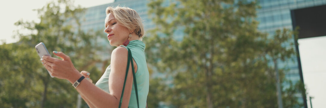 Friendly woman with short blonde hair in casual clothes looking at map trying to find his way using his mobile phone, panorama. Woman using map app in cellphone outdoors