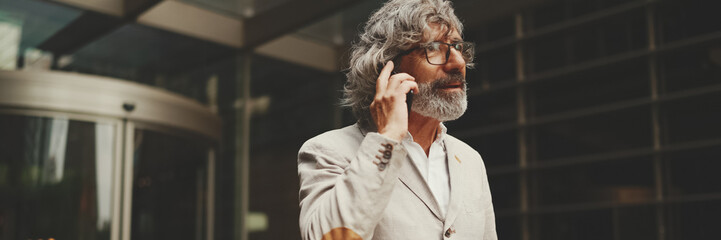 Mature businessman with beard and glasses wearing gray jacket walks up the stairs leaving the business center, Panorama. Middle aged manager talking on cellphone