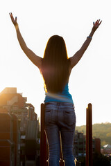 Portrait of a young attractive long haired woman in blue jeans with raised hands on the roof of building during sunset. Shot from behind. Beauty...