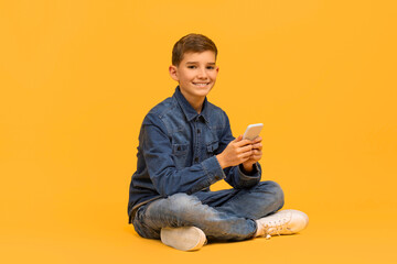 Smiling teen boy with smartphone in hands sitting cross-legged on yellow background