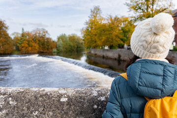 Unknown woman with a woolen hat and backpack enjoying beautiful views of the river from a stone bridge as it passes through a town in Ireland on an autumn day - obrazy, fototapety, plakaty