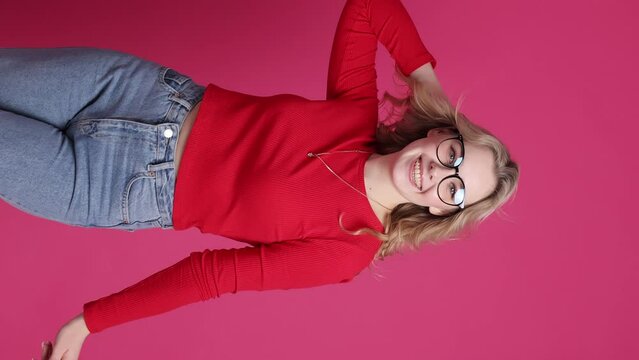 Cheerful young caucasian woman with transparent ceramic braces on teeth isolated on pink backdrop. Blond girl in eyeglasses looks at camera. Beauty, health and fashion concept. vertical video.
