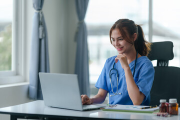 Friendly female nurse in blue scrubs seated at a desk, working on a laptop with medical charts and a stethoscope.. - obrazy, fototapety, plakaty