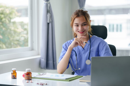 Friendly female nurse in blue scrubs seated at a desk, working on a laptop with medical charts and a stethoscope..