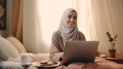 Beautiful and elegant millennial Asian Muslim woman or businesswoman with hijab using laptop computer to manage her tasks while relaxing on her comfortable sofa