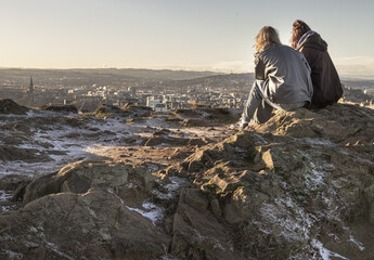 Two women sitting relaxing on top of rock over the mountain and enjoying looking at scenic view of...