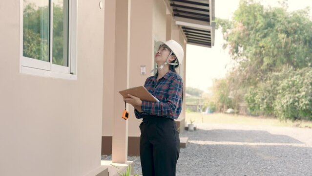 Professional Asian female inspector reviews construction details on clipboard while assessing home's exterior. Intently,  woman in safety helmet documents findings on structure of residential building