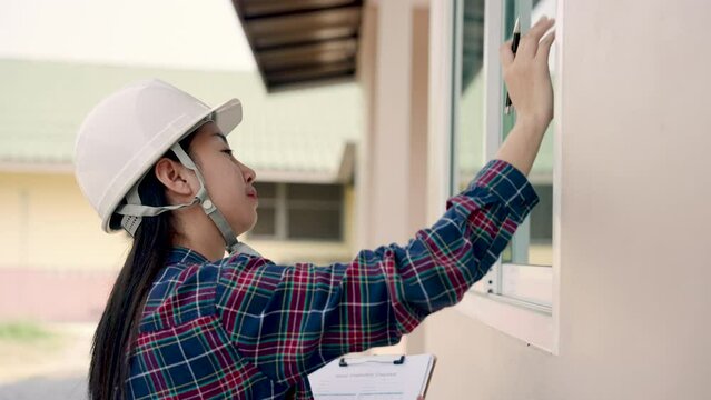 Asian female building inspector meticulously checks house exteriors, clipboard in hand, attentive woman in a safety helmet examines building details, pen poised for noting critical observations.