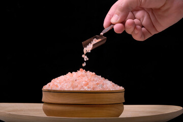 In his hand, spice spoon is pouring pink salt into cup on black background.