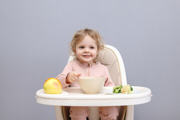 Cheerful little toddler baby girl eating healthy food while sitting in high chair looking at camera isolated over gray background having lunch in the morning at home