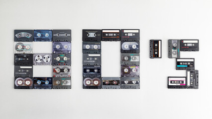The text "80's" on a gray paper background, composed of ancient, different, colorful audio cassettes. The year of nostalgia for analog home audio technology of the 1980s