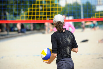 Beach volleyball. Young woman volley player standing in front of a net holding the ball waiting to toss it, blurred players on a background