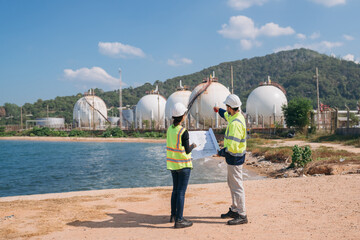 Two engineers in safety gear with blueprints discussing work in front of industrial storage tanks on a clear day.