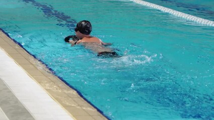 swimming pool training lesson for kids. little boy child learning to swim holding green floating board learning to move legs under water float on his side. child in swimming shorts, black cap and blue