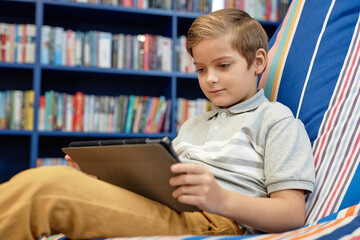 Portrait of young boy using digital tablet while relaxing in bean bag at modern library loungewith copy space