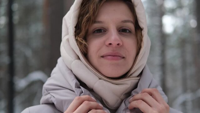 A young woman stands against a background of fir trees under a snowy forest in winter. A girl in a gray down jacket with a hood stands and looks at the camera. Close-up.