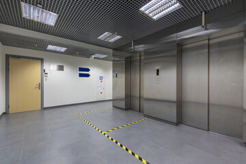 A lobby in a public building with two elevators. Ceiling lights and grey floor tiles.