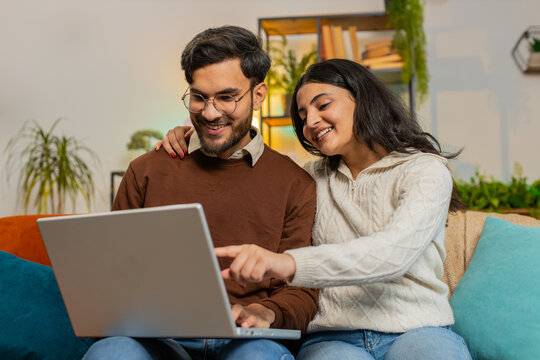 Cheerful young diverse couple in casual clothes discussing while using laptop on sofa in living room at home. Happy Indian family planning together pointing showing booking online tickets in apartment - Powered by Adobe