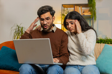 Shocked young diverse couple using laptop and covering mouth sitting on sofa in room at home. Upset disappointed Hispanic family man woman together reading bad news fail lose on netbook in apartment.