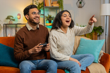 Cheerful young Indian couple using joystick controller playing video game fun enjoying sitting on...