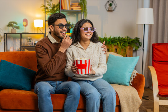 Cheerful young Arabian couple in 3D glasses eating popcorn and watching comedy movie sitting on sofa in living room at home. Smiling diverse family laughing enjoying film during weekend in apartment.