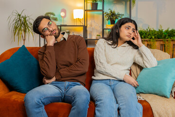 Upset stressed bored tired diverse family couple sitting on sofa at home room apartment. Man with...