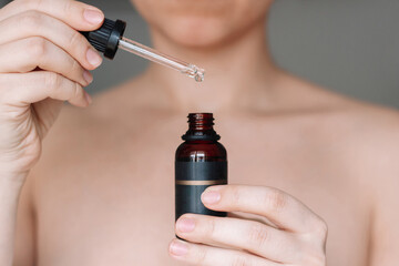 Cropped shot of a young woman holding a dropper and a glass bottle with essential cosmetic oil on a dark gray background. Skin care. Cosmetology concept