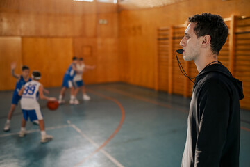 Side view of a trainer on court using whistle while boys practicing basketball.