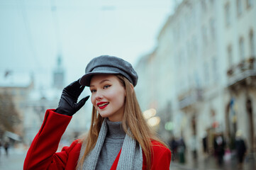 Fototapeta premium Fashionable happy smiling woman wearing elegant red coat, gray cap, black leather gloves, posing in street of European city. Outdoor close up portrait. Copy, empty, blank space for text 
