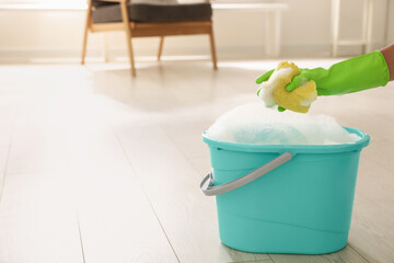 Woman holding sponge with foam over bucket indoors, closeup. Cleaning supplies © New Africa