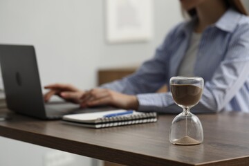 Hourglass with flowing sand on desk. Woman using laptop indoors, selective focus - Powered by Adobe